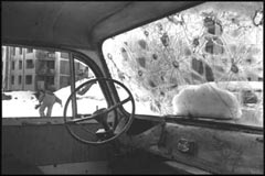 interior view of car with windows shot out, figure running towards burnt-out buildings in background