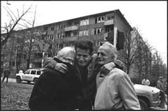 Young man with arms around shoulders of two old people in a park, a wrecked building looming behind them