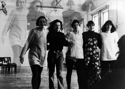 Five women standing in a row with arms around each other, their images cast on the wall behind them