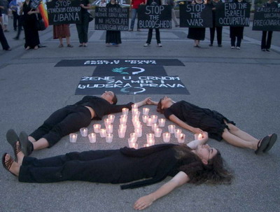 Three Women in Black form a triangle, lying on their backs on the pavement,  around lit candles arranged in a triangle