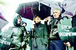 Jane and Eve wearing green raincoats under black umbrella, standing next to Eve's husband, holding green signs shaped like hands that say STOP THE OCCUPATION