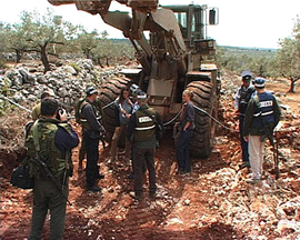 Neta and Yasmine chained to bulldozer, surrounded by soldiers, with olive grove and far hills around them